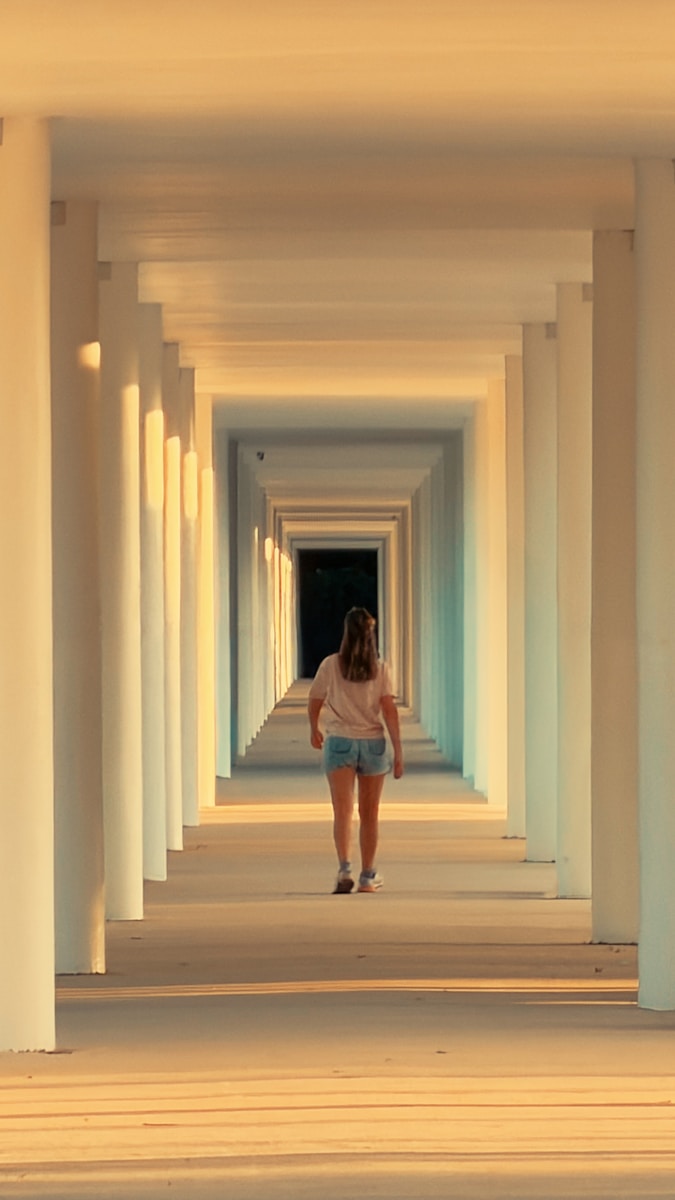 Photo by Milton Villemar Woman walks down a sunlit colonnade hallway.